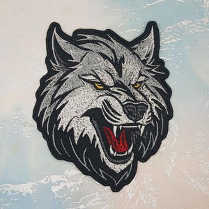 May include: Embroidered patch depicting a snarling wolf's head. The wolf has grey and white fur, yellow eyes, and a red mouth. The patch is outlined in black.