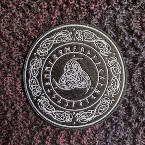 May include: Black and white embroidered patch with a circular design featuring a triskelion symbol surrounded by a circle of runes. The runes are in a circular pattern around the triskelion.