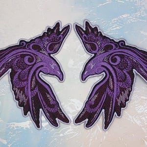 Könnte beinhalten: Zwei violette, gestickte Bügelbilder mit stilisierten Raben und filigranen Designs. Die Patches sind Spiegelbilder voneinander.