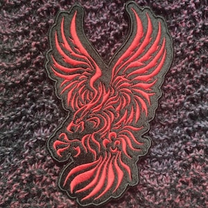 May include: A black and red embroidered patch of a stylized eagle with its wings spread wide. The eagle is outlined in black and filled in with red.
