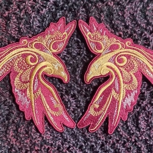 Könnte beinhalten: Zwei gestickte Bügelbilder in Form von stilisierten Phönixen mit goldfarbenem und rotem Garn. Die Phönixen sind einander zugewandt, mit ausgebreiteten Flügeln.