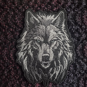 May include: A black and white embroidered wolf's head patch. The wolf has a fierce expression with sharp teeth and piercing eyes.