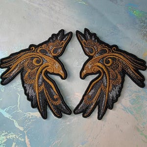 Könnte beinhalten: Zwei schwarze, gestickte Bügelbilder von stilisierten Raben mit goldenen Akzenten. Die Raben sind einander zugewandt und haben ihre Flügel ausgebreitet.