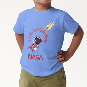 May include: A light blue t-shirt with a cartoon astronaut flying with a red cape and the text "I like to visit NASA" in a circle around the astronaut. The text "NASA" is printed in red below the astronaut.