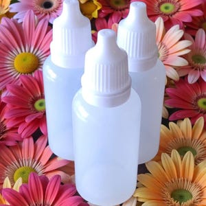 May include: Three clear plastic dropper bottles with white caps are set against a backdrop of colourful gerbera daisies. The flowers are in shades of pink, orange, and yellow, creating a vibrant and natural setting.