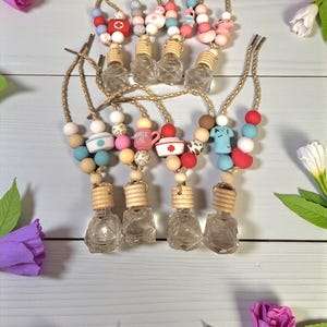 Puede incluir: Varias botellas pequeñas de vidrio con tapas de madera están suspendidas por cuerdas color beige. Cada cuerda tiene una serie de cuentas coloridas. Algunas cuentas tienen símbolos médicos y el texto "Nurse Life".