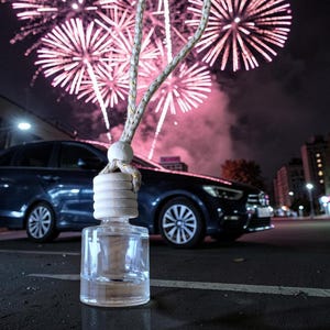 Puede incluir: Un ambientador de coche de vidrio transparente con una tapa de madera y una cuerda para colgar. El fondo presenta un coche azul oscuro y fuegos artificiales rosas explotando en el cielo nocturno. El coche está estacionado sobre asfalto oscuro.