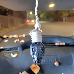 May include: A dark grey skull-shaped car air freshener suspended from a white and gold string. The air freshener has a white top and is hanging in front of a car dashboard. The background is blurred, showing a street scene.