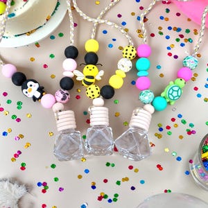 Op de afbeelding: Drie heldere glazen auto luchtverfrisser flesjes met houten doppen worden getoond. Elk flesje is bevestigd aan een kralenketting met kleurrijke kralen en bedels, waaronder een pinguïn, een bij en een schildpad. De achtergrond bevat confetti en ballonnen.