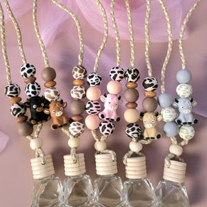 Op de afbeelding: Een verzameling auto-luchtverfrissers met decoratieve kralen en koeienbeeldjes. Elke verfrisser heeft een kleine glazen fles met een houten dop en een reeks kralen in verschillende kleuren en patronen, waaronder zwart-witte vlekken.