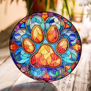 Peut inclure: Une fenêtre en verre coloré suspendue avec un motif de patte. L'empreinte de patte est composée de morceaux de verre rouge, orange, jaune, bleu et vert. La fenêtre est entourée d'une bordure décorative en verre bleu, violet et rouge.