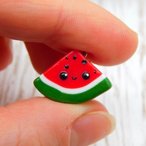 Könnte beinhalten: Ein kleiner, roter Wassermelonen-Schnitz-Anhänger mit weißer Schale und grünen Rändern. Die Wassermelone hat ein süßes Comic-Gesicht mit schwarzen Augen und einem Lächeln.