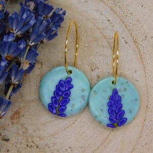 Könnte beinhalten: Goldene Creolen-Ohrringe mit runden, hellblauen Anhängern mit einem lila Lavendel-Design. Ein Bündel blauer Lavendelblüten befindet sich im Hintergrund, vor einem hellbraunen Untergrund.