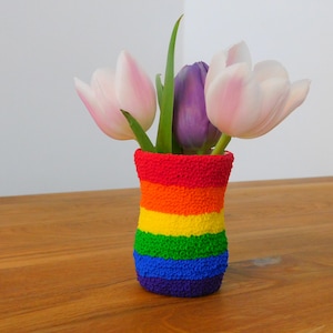 Könnte beinhalten: Eine regenbogenfarbene gehäkelte Vase mit drei rosa und lila Tulpen.