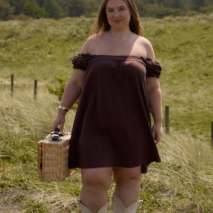 May include: A woman wearing a brown off-the-shoulder dress and cream-colored cowboy boots stands in a grassy field. She carries a wicker picnic basket. The dress has short sleeves. The background features green grass and trees.
