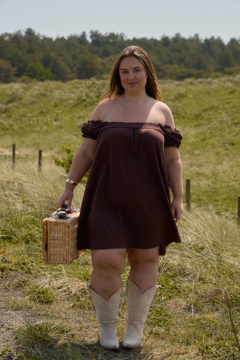 May include: A woman wearing a brown off-the-shoulder dress and cream-colored cowboy boots stands in a grassy field. She carries a wicker picnic basket. The dress has short sleeves. The background features green grass and trees.
