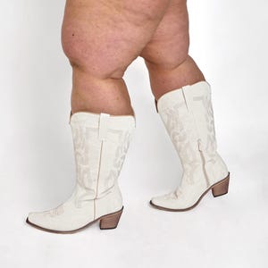 Op de afbeelding: Off-white cowboylaarzen met ingewikkelde geborduurde patronen. De laarzen hebben een puntige neus, een zijrits en een bruine blokhak. De laarzen zijn kniehoog en gemaakt van een zacht materiaal.