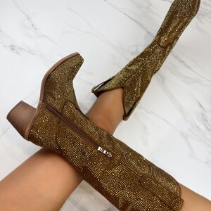 May include: A pair of gold-colored cowboy boots covered in small, sparkling embellishments. The boots have a pointed toe, a side zipper, and a stacked heel. The boots are knee-high and have a classic Western design.
