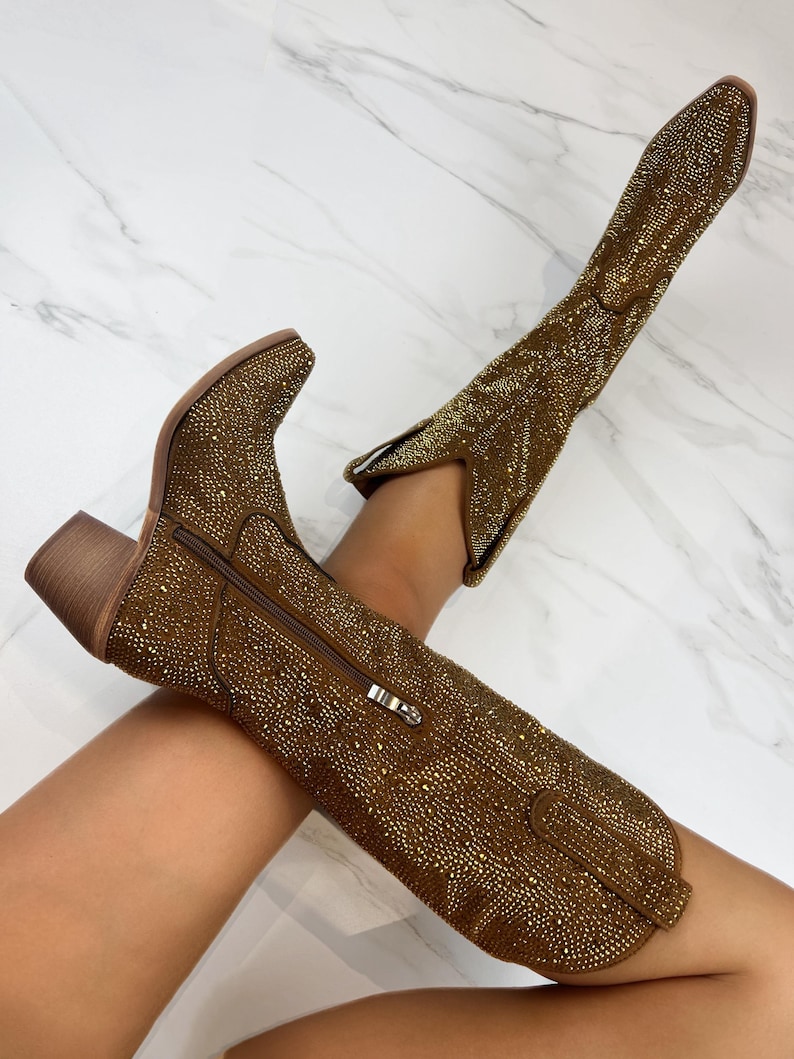 May include: A pair of gold-colored cowboy boots covered in small, sparkling embellishments. The boots have a pointed toe, a side zipper, and a stacked heel. The boots are knee-high and have a classic Western design.
