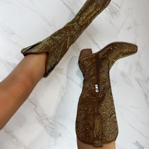 May include: Two brown knee-high boots covered in small, shiny gold embellishments. One boot is upright, while the other is angled. The boots have pointed toes, short heels, and zippers on the sides.