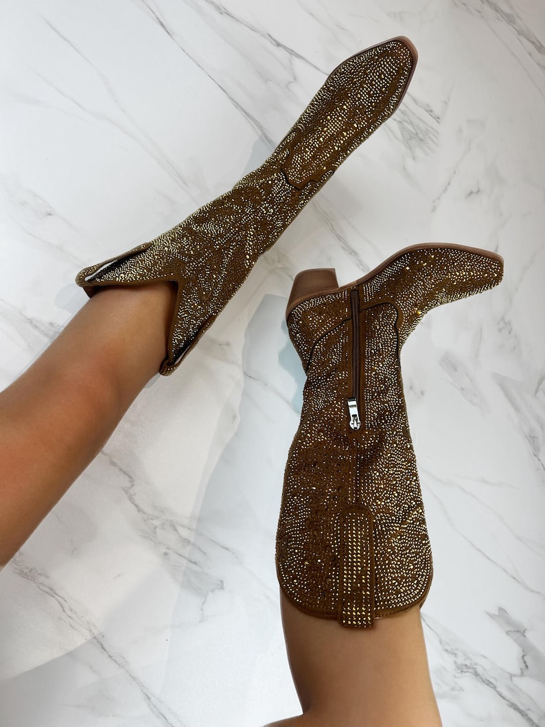 May include: Two brown knee-high boots covered in small, shiny gold embellishments. One boot is upright, while the other is angled. The boots have pointed toes, short heels, and zippers on the sides.
