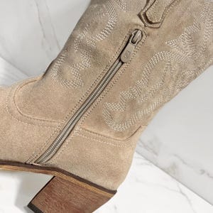 May include: A close-up of a beige suede cowboy boot with intricate stitched patterns. The boot features a side zipper and a stacked wooden heel. The stitching creates a decorative design on the boot's shaft.