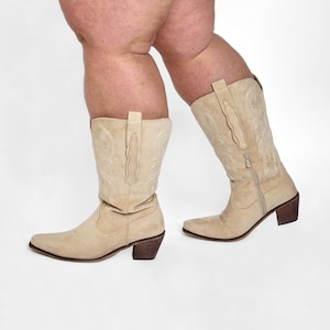 May include: A pair of beige suede cowboy boots with pointed toes and stacked heels. The boots have decorative stitching and a zipper on the side. The boots are shown against a white background.