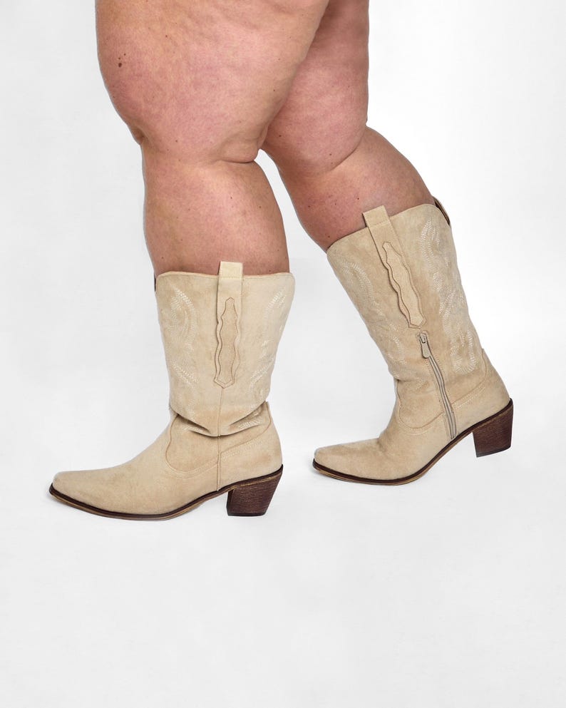 May include: A pair of beige suede cowboy boots with pointed toes and stacked heels. The boots have decorative stitching and a zipper on the side. The boots are shown against a white background.