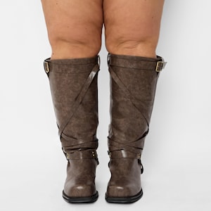 Könnte beinhalten: Braune kniehohe Stiefel mit dekorativen Riemen und Schnallen. Die Stiefel haben ein strukturiertes Aussehen und einen niedrigen Absatz. Die Riemen kreuzen sich auf der Vorderseite der Stiefel und sorgen für ein stilvolles Detail. Die Stiefel sind aus Kunstleder.