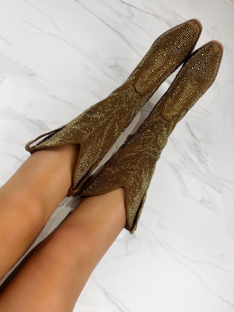 May include: A pair of gold-colored boots with a pointed toe and a low heel. The boots are covered in small, sparkling embellishments, giving them a glamorous appearance. The boots are angled towards the viewer, showcasing their design and texture.