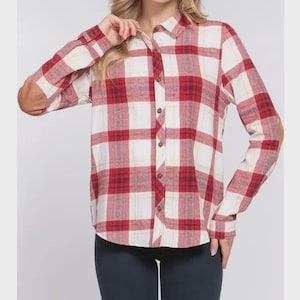 Puede incluir: Una camisa a cuadros roja, blanca y crema con mangas largas y coderas. La camisa tiene un cuello clásico y botones de madera. La modelo lleva leggings azul oscuro.