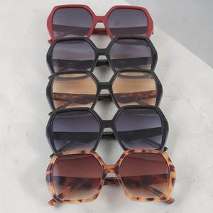 May include: Five pairs of oversized sunglasses in a row. The top pair is red, followed by black, tortoise shell, dark grey, and a second tortoise shell pair at the bottom. All have an octagonal, geometric shape.