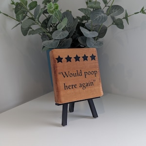 May include: A square wooden sign with a light brown finish, featuring five black stars and the black text "Would poop here again" printed on the front. The sign is displayed on a small black tripod stand, positioned in front of a green plant.