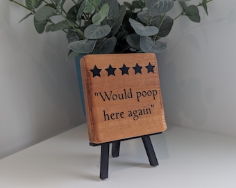 May include: A square wooden sign with a light brown finish, featuring five black stars and the black text "Would poop here again" printed on the front. The sign is displayed on a small black tripod stand, positioned in front of a green plant.