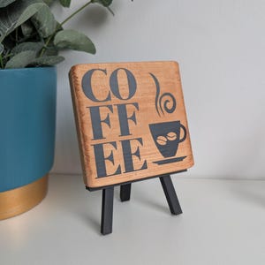 May include: A square wooden sign with a natural light brown finish, featuring the black text COFFEE stacked vertically and a graphic of a steaming cup with two coffee beans. The sign is displayed on a small black metal tripod easel stand.