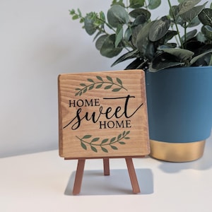 May include: A square wooden sign featuring the text "HOME sweet HOME" in black lettering, accented by green leaf illustrations above and below the text. The sign is displayed on a small, three-legged brown easel stand.
