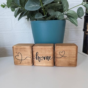 May include: Three wooden blocks spelling out the word "home". Each block features a small heart design. The blocks are displayed on a white surface.