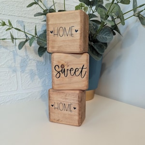 May include: Three stacked light brown wooden blocks with a natural grain finish. The top and bottom blocks feature the word HOME in black capital letters with small hearts on each side. The middle block features the word sweet in black script.