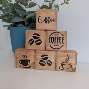May include: Six wooden blocks stacked, each with a coffee theme. The blocks display coffee beans, cups, and the word "Coffee". The blocks are made of light-coloured wood, and the designs are in black.