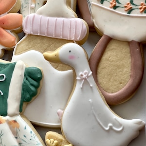 May include: A set of five decorated sugar cookies, including a baby bottle, a rattle, a duck, and two floral designs. The cookies are decorated with white, pink, green, and brown icing.