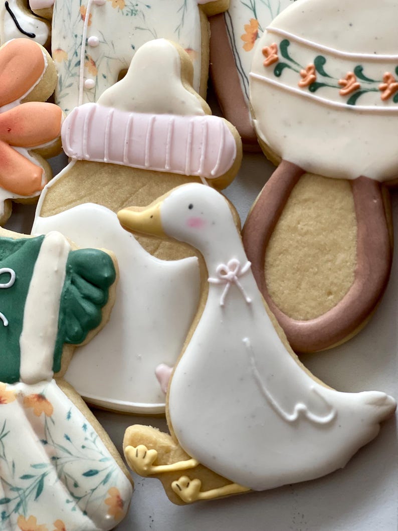 May include: A set of five decorated sugar cookies, including a baby bottle, a rattle, a duck, and two floral designs. The cookies are decorated with white, pink, green, and brown icing.