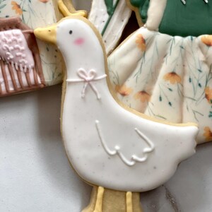 May include: A white goose cookie with yellow feet and a pink bow. The cookie is decorated with white icing and has a floral pattern.