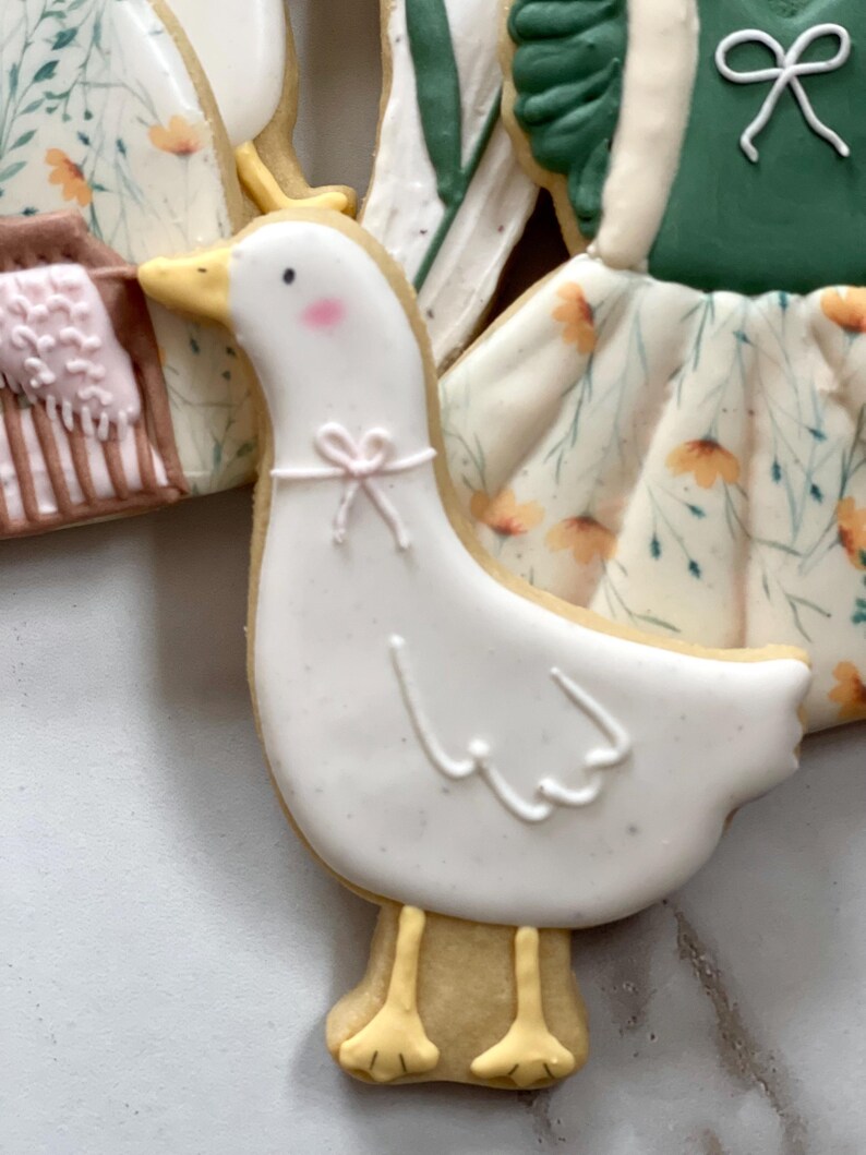 May include: A white goose cookie with yellow feet and a pink bow. The cookie is decorated with white icing and has a floral pattern.