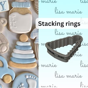 May include: A set of cookie cutters for making baby-themed cookies. The cutters are made of metal and are shaped like a baby bottle, a stacking ring toy, a car, and a footprint. The cutters are shown next to cookies that have been cut out using the cutters.