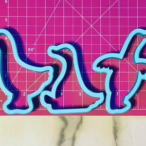 May include: Three blue plastic cookie cutters shaped like geese. The cutters are on a pink cutting mat with white grid lines.