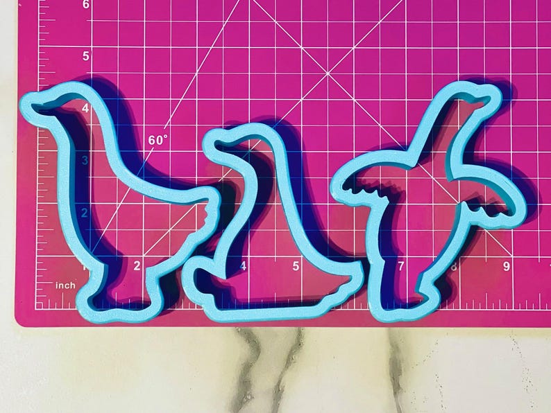 May include: Three blue plastic cookie cutters shaped like geese. The cutters are on a pink cutting mat with white grid lines.