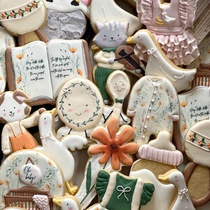 May include: A collection of decorated sugar cookies featuring a variety of baby-themed designs, including a cow, a goose, a flower, a book, a rattle, a spoon, a crib, and a dress. The cookies are decorated with pastel colors and floral patterns.