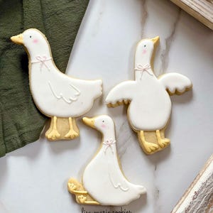 May include: Three white sugar cookies decorated with yellow and pink icing in the shape of geese. The cookies are on a white marble surface. The text "lisa marie cookies" is written in black on the surface.