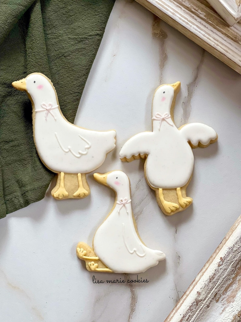 May include: Three white sugar cookies decorated with yellow and pink icing in the shape of geese. The cookies are on a white marble surface. The text "lisa marie cookies" is written in black on the surface.