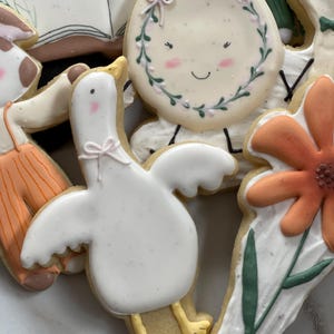 May include: A set of decorated sugar cookies featuring a white goose with yellow feet, a flower with orange petals, a book with the text "the cow jumped over the moon", and a round cookie with a wreath and a smiling face.